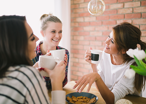 Three People Sharing Coffee Around A Wooden Table With Cookies And Tulips, Set In A Cozy Indoor Space.