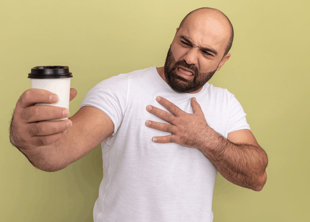 A Man In A White T-Shirt Holds A Disposable Coffee Cup With A Black Lid, Grimacing As He Places One Hand On His Chest. His Expression Conveys Discomfort And Concern, As If He'S Questioning How Does Coffee Affects Heart Health.